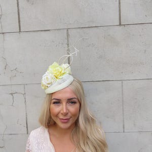 Pode incluir: Um chapéu de flores branco e amarelo com uma base redonda pequena e uma única pena branca. O chapéu é usado por uma pessoa com cabelo comprido e loiro.