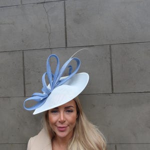 May include: A formal headpiece featuring a wide, flat, white circular base adorned with a series of upright, looped light blue ribbons. The structure includes a single thin, curved white quill extending upward from the center of the blue loops.
