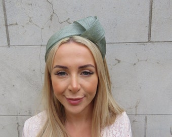 May include: A sage green fabric headband with a knotted design. The headband is worn by a person with long blonde hair.