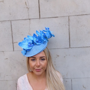 May include: A light blue hat with a large blue orchid flower embellishment. The hat is made of a soft, textured fabric and has a wide brim.