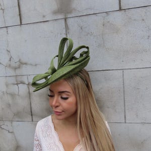 May include: A green headpiece with a looped design. The headpiece is worn on the head. The woman is wearing a white lace top. The background is a light gray stone wall.