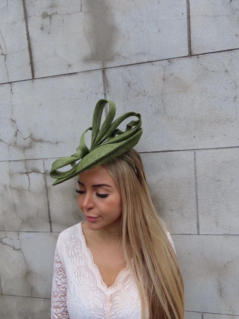 May include: A green headpiece with a looped design. The headpiece is worn on the head. The woman is wearing a white lace top. The background is a light gray stone wall.
