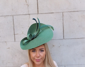 May include: A vibrant emerald green fascinator hat featuring a wide, circular brim with a coiled straw texture. The hat is adorned with a decorative loop of matching green straw material and several iridescent peacock feathers extending upwards.