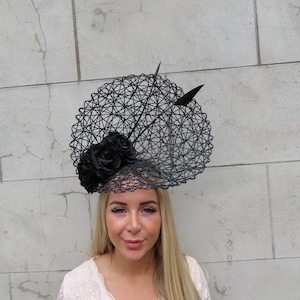 Puede incluir: Un fascinador negro de tejido abierto con un diseño circular, adornado con rosas de tela negra y dos acentos de plumas negras. El tocado está diseñado para ser usado en la cabeza.