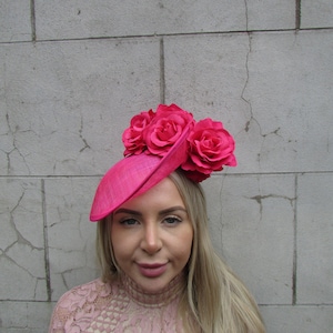 May include: A bright pink hat with three large pink fabric roses on top. The hat is tilted to the side and the woman wearing it has blonde hair.