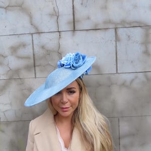 May include: A light blue, wide-brimmed hat with a cluster of matching blue fabric roses. The hat is worn with a beige coat and a light-colored top. The background is a textured, light gray wall.