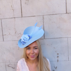 May include: A light blue, wide-brimmed hat with a decorative floral arrangement and two feathers. The hat features a large, light blue rose and smaller blue flowers. This elegant hat is suitable for special occasions.
