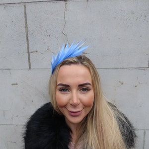 May include: A light blue feather headband with a pointed design. The headband is worn by a person with long blonde hair.