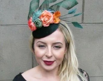 May include: A black fascinator hat featuring a cluster of peach, orange, and green fabric roses, accented with tall, structured dark green mesh loops and thin, pointed dark green feathers extending outward from the side of the base.