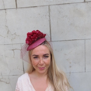 Könnte beinhalten: Ein dunkelroter Fascinator mit einer runden Basis, einem Arrangement aus passenden Stoffblumen und einem feinen Netzschleier, der über die Vorderseite fällt. Das Kopfstück wird schräg getragen und zeigt die strukturierte Oberfläche und dunkle Töne.