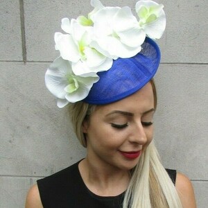 Op de afbeelding: Een koningsblauwe fascinator hoed versierd met grote witte orchidee bloemen. De hoed heeft een cirkelvormige basis en is versierd met verschillende grote witte orchidee bloemen met gele centra. De hoed wordt gedragen door een persoon met blond haar en een zwarte top.