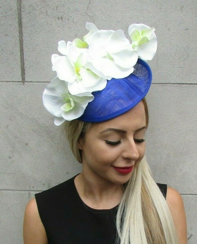 Op de afbeelding: Een koningsblauwe fascinator hoed versierd met grote witte orchidee bloemen. De hoed heeft een cirkelvormige basis en is versierd met verschillende grote witte orchidee bloemen met gele centra. De hoed wordt gedragen door een persoon met blond haar en een zwarte top.