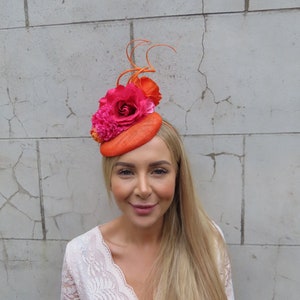 Könnte beinhalten: Ein leuchtend orangefarbener Fascinator mit einem Blumenarrangement in Pink und Orange. Der Fascinator ist aus Stroh gefertigt und hat einen kleinen Rand.