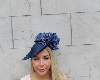 May include: A navy blue hat with a wide brim and a cluster of blue fabric flowers. The hat is worn by a person with long blonde hair and a light-colored lace top. The background is a light gray brick wall.