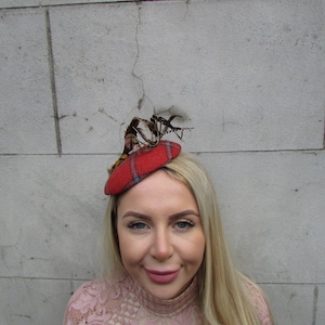 以下が含まれることがあります： ピンクのレースのトップスを着た女性が、小さな赤と黒の格子柄の帽子をかぶっています。帽子の上には羽根が付いています。