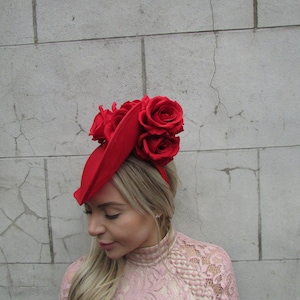 Könnte beinhalten: Ein leuchtend roter Fascinator mit roten Rosen. Der Kopfschmuck wird auf dem Kopf getragen. Die Person trägt ein rosa Spitzenkleid. Der Hintergrund ist eine graue Backsteinmauer.