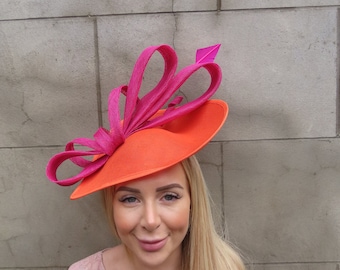 Könnte beinhalten: Ein leuchtend orangefarbener Hut mit einer großen rosa Schleife und einer einzelnen rosa Feder. Der Hut wird von einer Person mit langen blonden Haaren getragen.