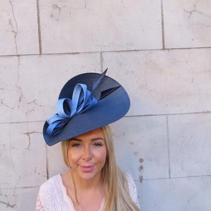 May include: A navy blue hat with a wide brim, adorned with a large, layered blue bow and a black feather. The hat is a stylish accessory, perfect for special occasions.