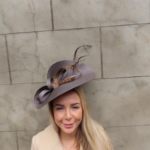 Puede incluir: Un sombrero gris con ala ancha, adornado con un lazo gris oscuro y plumas. Este elegante sombrero es un accesorio de moda, ideal para ocasiones especiales. El sombrero lo lleva una persona con un abrigo beige.