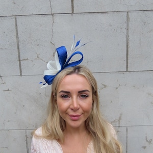 Pode incluir: Um fascinator azul e branco com detalhes de penas. O adorno de cabeça apresenta um grande laço azul e detalhes brancos. O fascinator é usado na cabeça. O fundo é uma parede de tijolos cinza.