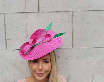 May include: A pink hat with green accents. The hat is made of straw and has a wide brim. The hat is decorated with a green bow and green feathers.