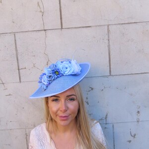May include: A light blue hat with a wide brim, adorned with clusters of blue fabric flowers. The hat has a feather accent and is likely made of a woven material. The hat is a stylish accessory.