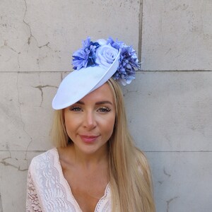 May include: A light blue hat with a large flower embellishment. The flower is made of blue and white fabric and is centered on the hat. The hat is tilted slightly to the side.