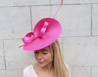 May include: A bright pink, wide-brimmed hat with a large bow on the side. The hat is made of a straw-like material and has a long, thin feather extending from the back.