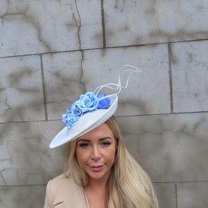 May include: A white hat with a wide brim, adorned with light blue fabric roses and a delicate, swirling accent. The hat is designed for special occasions and events.