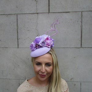 May include: A lavender hat with a small brim and a cluster of purple and pink flowers on top. The hat is decorated with a long, thin, curled purple feather.