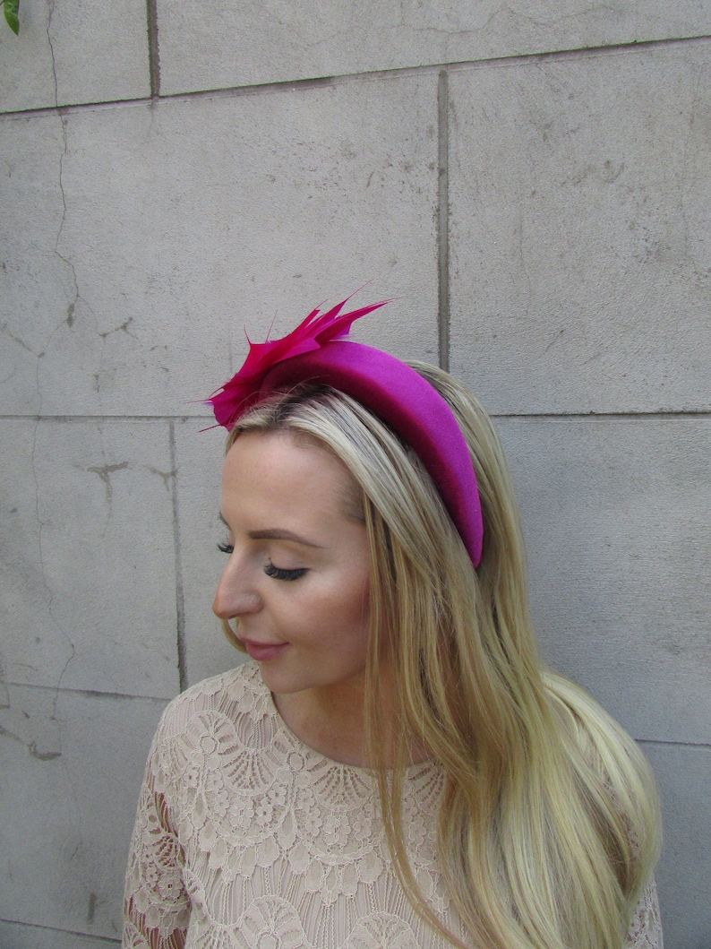 May include: A bright pink velvet headband with a matching pink feather accent. The headband is worn on a person with long blonde hair.