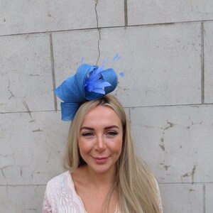 May include: A blue headpiece with a woven texture, featuring a large bow and delicate feather accents. The headpiece is a vibrant shade of blue, suitable for special occasions. The design includes a rolled brim and a decorative arrangement of feathers.