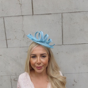 Pode incluir: Um fascinator azul claro com um design encaracolado. O fascinator é feito de palha e tem uma pequena base redonda.