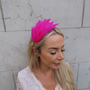 May include: A bright pink feather headband with a wide, flat band and a large, dramatic arrangement of feathers. The headband is worn by a person with long, blonde hair.