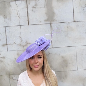 May include: A lavender-colored hat with a wide brim and a floral accent. The hat is worn by a person with long blonde hair and a white lace top. The background is a light-colored brick wall.