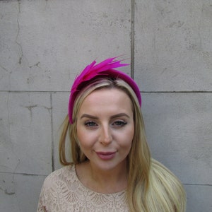 May include: A bright pink headband with a large feather accent. The headband is worn by a person with long blonde hair.