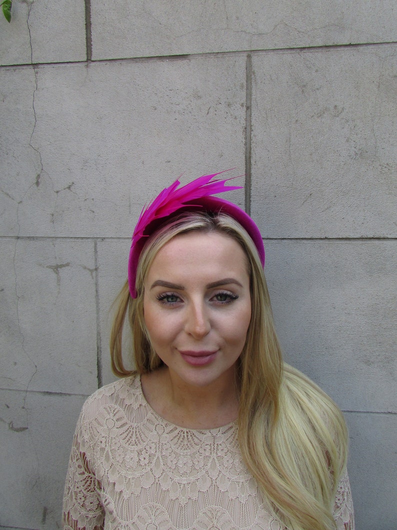 May include: A bright pink headband with a large feather accent. The headband is worn by a person with long blonde hair.