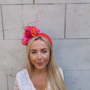 May include: A bright pink headband with a large cluster of pink, orange, and white flowers. The headband has a long, thin pink feather extending from the flower cluster.