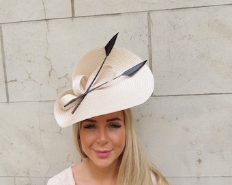 May include: A cream-colored, wide-brimmed hat with black decorative accents. The hat features a woven texture and is adorned with two black, stylized arrow-shaped embellishments. The hat is a stylish accessory.