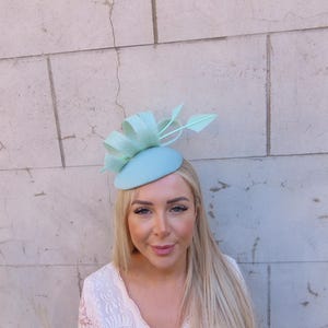 Könnte beinhalten: Hellblauer Fascinator-Hut mit Schleife und Blättern. Der Hut ist hellblau und aus glattem Material gefertigt. Der Hut wird auf dem Kopf getragen.