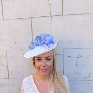 May include: A white hat with a wide brim, adorned with a light blue ribbon and a cluster of blue and purple flowers. The hat is a stylish accessory, perfect for special occasions.
