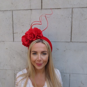 Op de afbeelding: Een rood hoofdband met een grote rode bloemenaccent en een lang, dun rood lint dat omhoog reikt. Het hoofdband wordt gedragen door een persoon met lang blond haar.