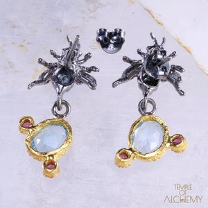 May include: A pair of silver and gold earrings with a blue gemstone and pink accents. The earrings feature a detailed design of a stylized insect with multiple legs.