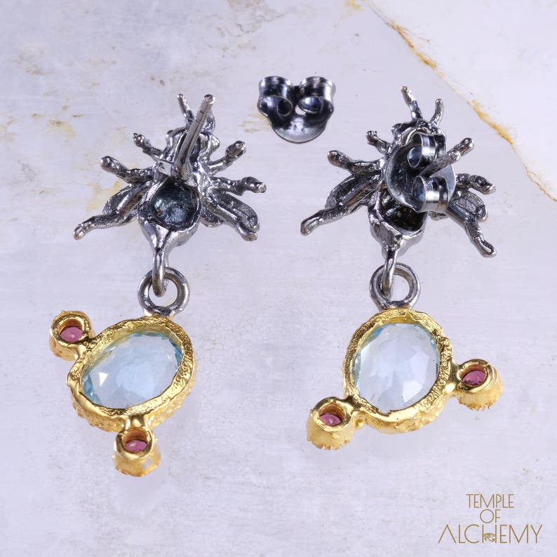 May include: A pair of silver and gold earrings with a blue gemstone and pink accents. The earrings feature a detailed design of a stylized insect with multiple legs.