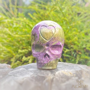 Può includere: Un teschio di cristallo verde e viola con un cuore intagliato sulla fronte. Il teschio è seduto su una roccia.