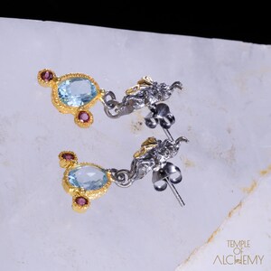 May include: A pair of silver and gold earrings with a blue gemstone and pink accents. The earrings feature a delicate, intricate design.