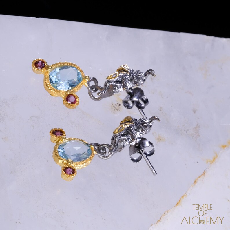 May include: A pair of silver and gold earrings with a blue gemstone and pink accents. The earrings feature a delicate, intricate design.