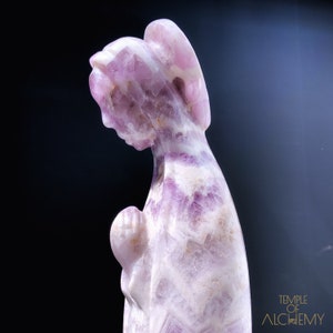 May include: A carved amethyst statue of a woman with wings, praying with her hands clasped together. The statue is a light purple color with white streaks throughout.