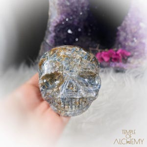 May include: A carved crystal skull with blue, brown, and clear tones. The skull is held in a hand, with a blurred background featuring purple crystals and pink flowers. The skull is a decorative item.
