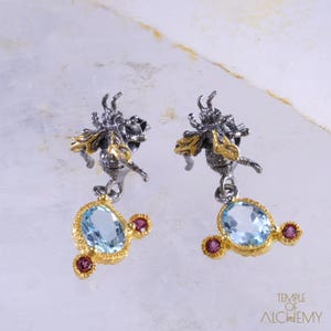 May include: A pair of silver and gold earrings featuring a bee design with a dangling blue gemstone and red accents. The earrings are a statement piece with a unique and intricate design.